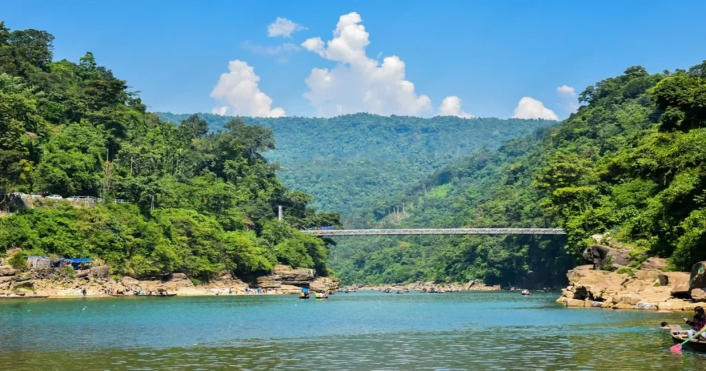 A serene river flows through a lush green valley in Jaflong, with a metal bridge spanning over it. The landscape is framed by dense forests on hillsides under a clear blue sky with scattered clouds. Several boats and people enjoy the tranquil setting.