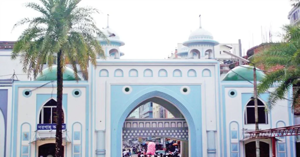 Historic blue and white gateway with arches and domed towers at the Shrine of Hazrat Shah Jalal, standing against a clear sky. A tall palm tree is on the left, and people walk underneath near shops in the background.