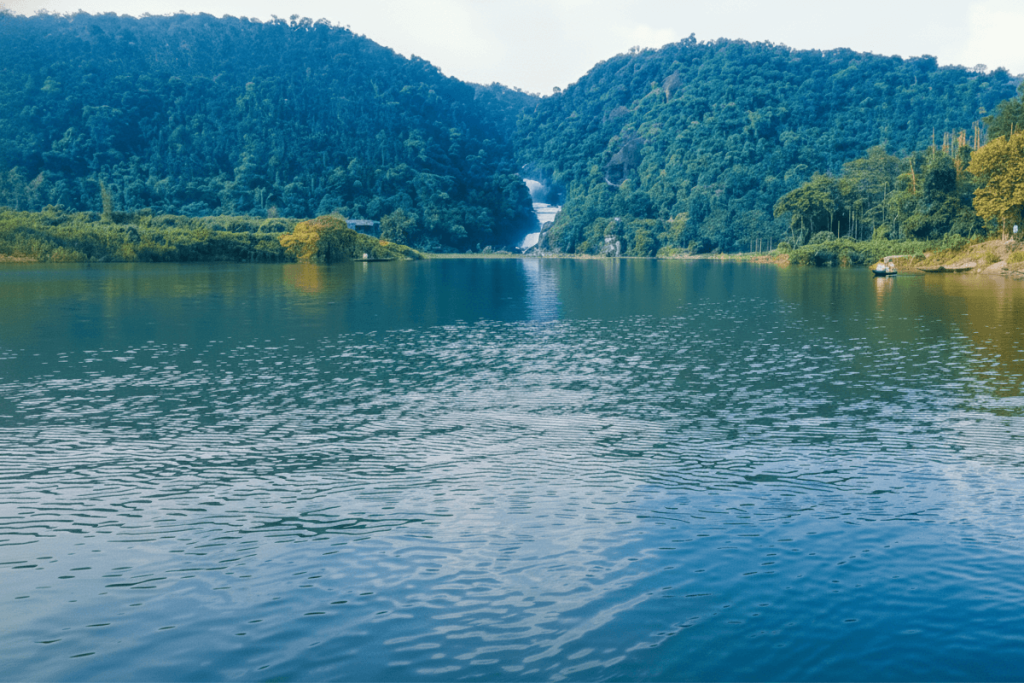 Panthumai Waterfall Village Viewpoint, one of Sylhet's Hidden Gems, showing a tranquil village field on the Bangladesh side with a massive, multi-tiered waterfall cascading down the green hills across the border in the distance.